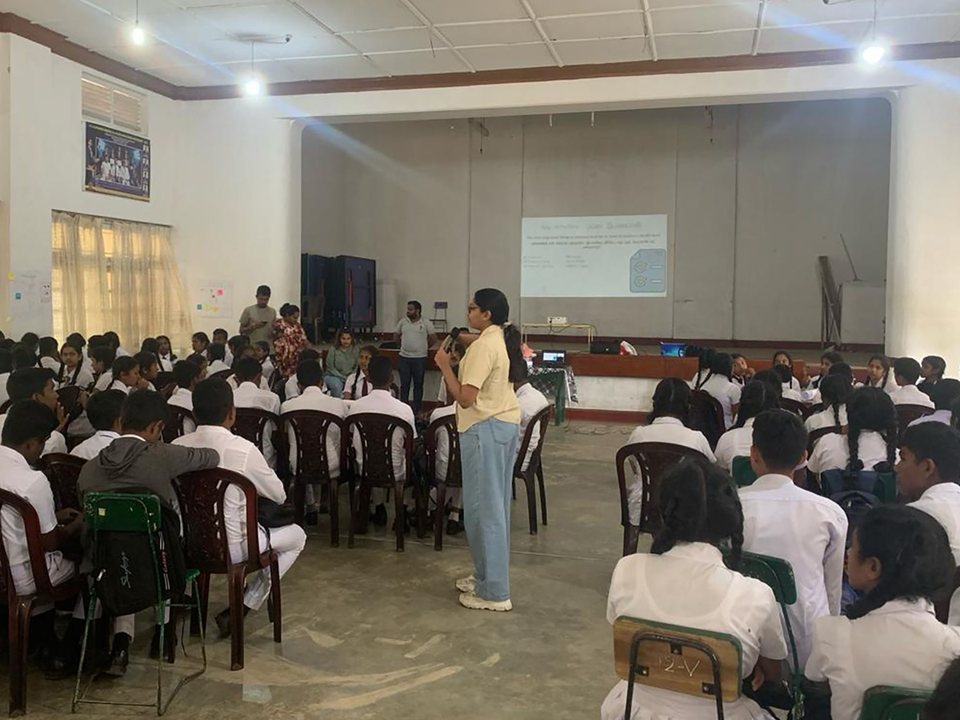 Chamathka speaking with a microphone to a hall of school students